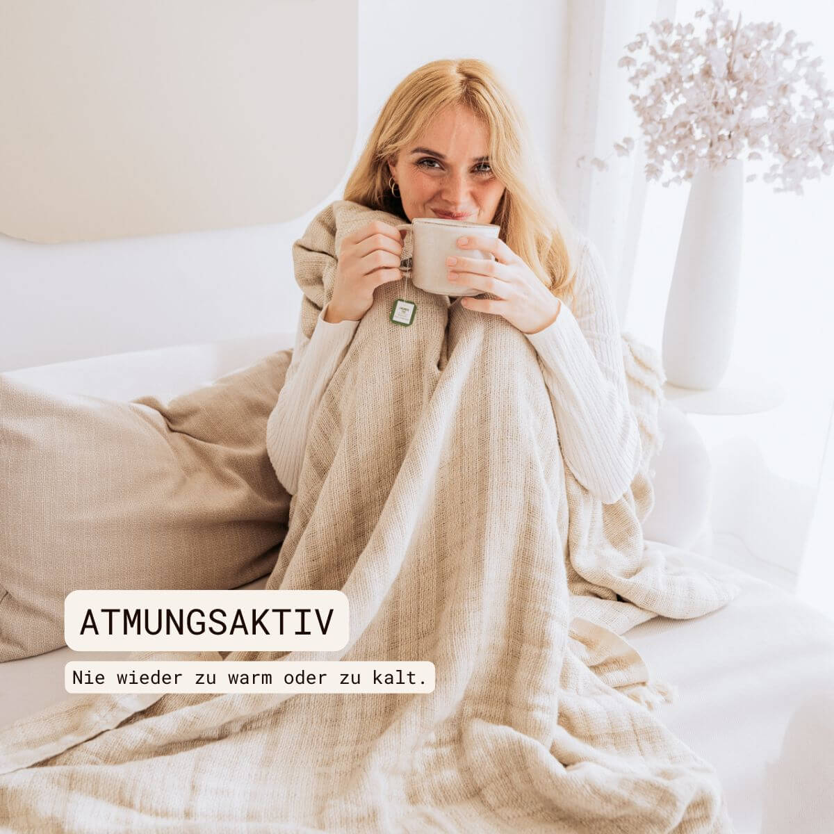 Kuscheldecke (135x200 cm) aus 70% Leinen und 30% OEKO-TEX zertifizierter Baumwolle | Kuscheldecke in der Farbe Beige | Frau sitzt eingewickelt in beigefarbene Decke im Wohnzimmer | Atmungsaktiv