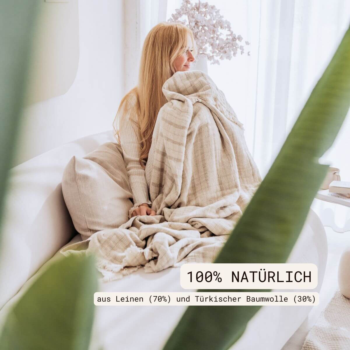 Kuscheldecke (135x200 cm) aus 70% Leinen und 30% OEKO-TEX zertifizierter Baumwolle | Kuscheldecke in der Farbe Beige | Frau sitzt mit Decke auf der Couch | 100% natürlich