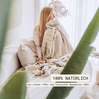 Kuscheldecke (135x200 cm) aus 70% Leinen und 30% OEKO-TEX zertifizierter Baumwolle | Kuscheldecke in der Farbe Beige | Frau sitzt mit Decke auf der Couch | 100% natürlich