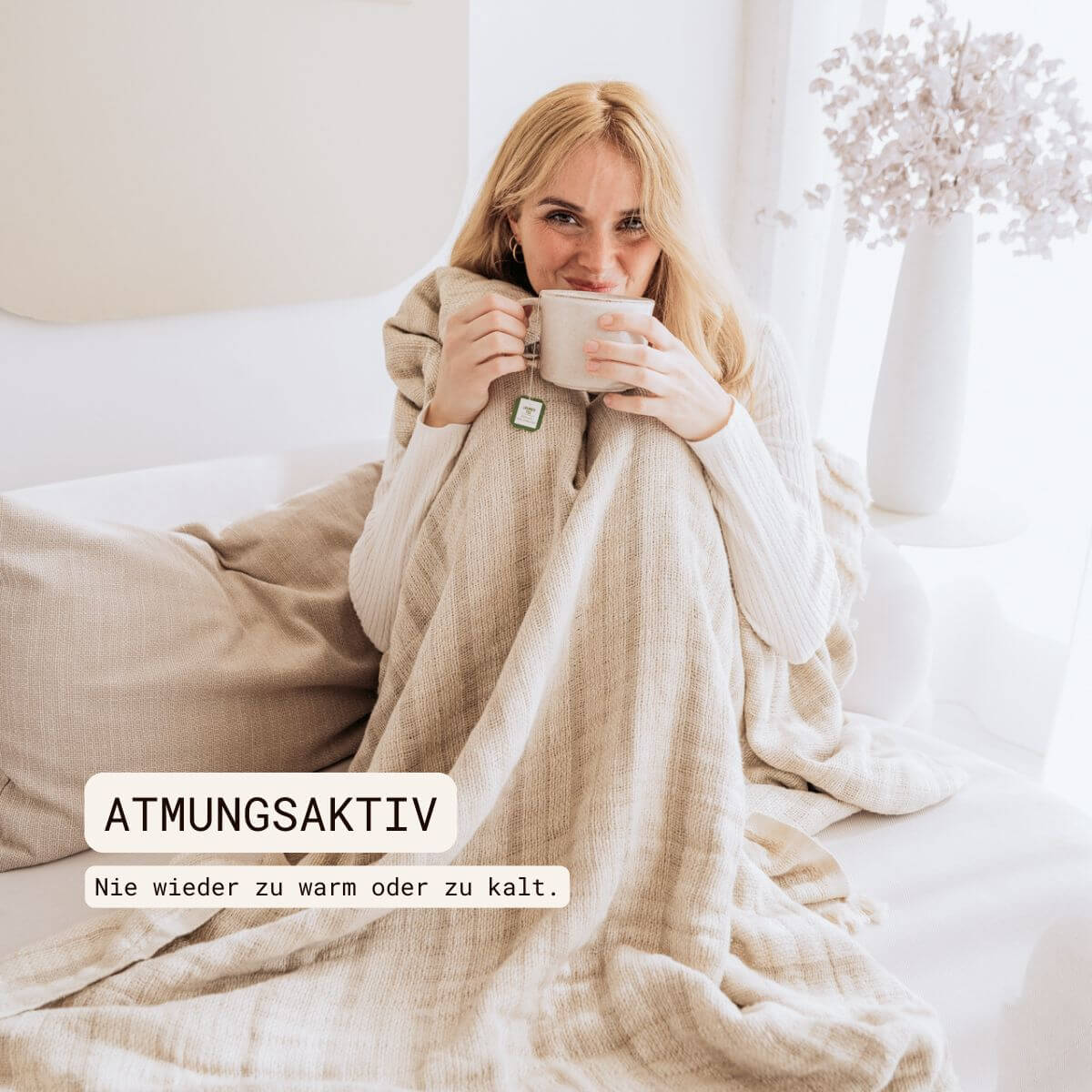 Kuscheldecke (135x200 cm) aus 70% Leinen und 30% OEKO-TEX zertifizierter Baumwolle | Kuscheldecke in der Farbe Beige | Frau sitzt eingewickelt in beigefarbene Decke im Wohnzimmer | Atmungsaktiv