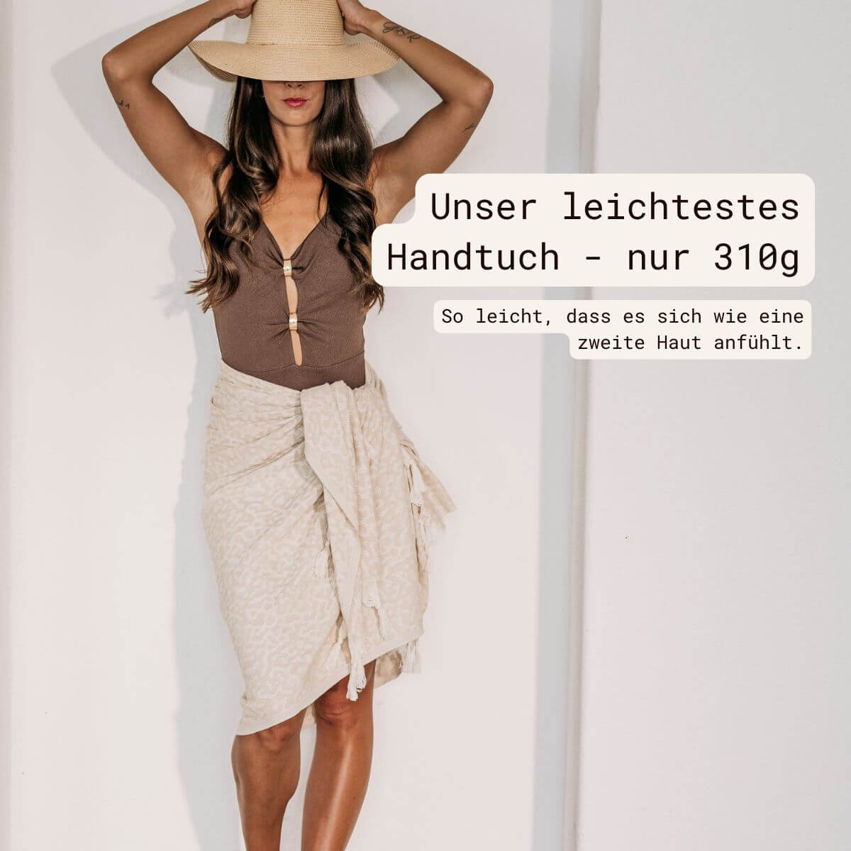 Frau mit beigem VAYVA Strandtuch als Sarong um die Hüften gewickelt, dazu brauner Badeanzug und Strohhut