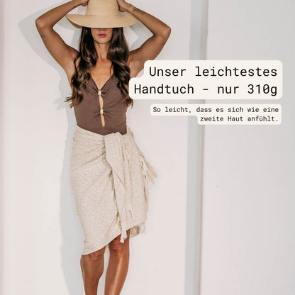 Frau mit beigem VAYVA Strandtuch als Sarong um die Hüften gewickelt, dazu brauner Badeanzug und Strohhut